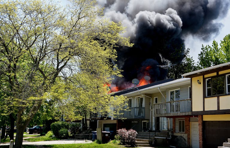 Dramatic house fire with thick smoke rising in a suburban neighborhood, surrounded by trees.